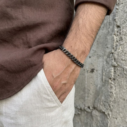A close-up of a man's wrist wearing a dark metallic chain link bracelet, styled with a brown linen shirt and off-white linen trousers against a textured concrete wall.