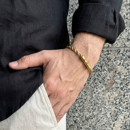 A close-up of a person's hand wearing a gold-colored twisted rope bracelet with a sliding clasp, styled with a black shirt and light beige trousers against a gray stone background.