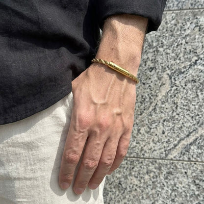 A close-up of a person's hand wearing a gold-toned twisted rope bracelet with a polished cylindrical bar, set against a gray stone background.