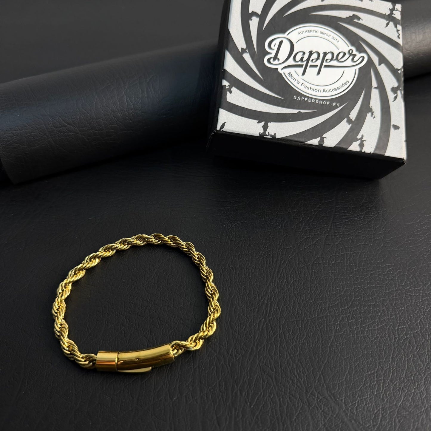 A gold-colored twisted rope chain bracelet with a polished cylindrical clasp, displayed on a black textured surface next to its black and white branded Dapper jewelry box.