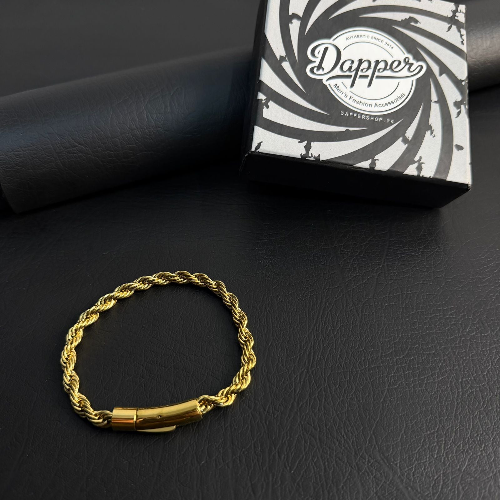 A gold-colored twisted rope chain bracelet with a polished cylindrical clasp, displayed on a black textured surface next to its black and white branded Dapper jewelry box.