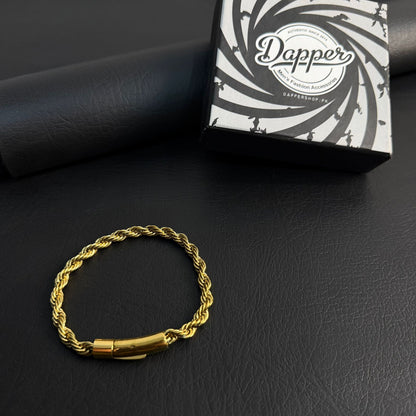 A gold-colored twisted rope chain bracelet with a polished cylindrical clasp, displayed on a black textured surface next to its black and white branded Dapper jewelry box.