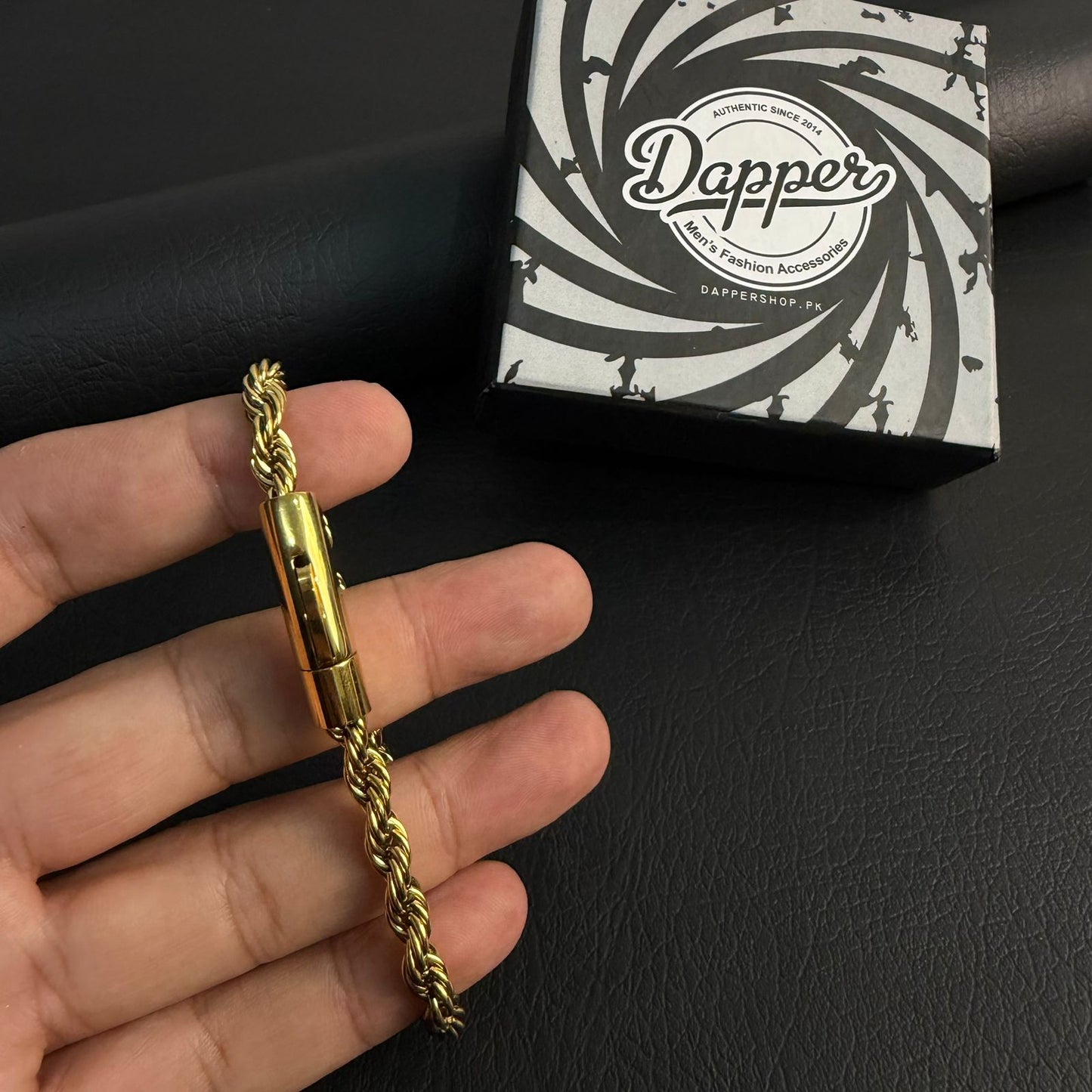 A gold-toned rope chain bracelet with a toggle clasp held in a hand, with its branded black and white box in the background.