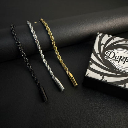 Three rope-style bracelets in black, silver, and gold are displayed on a dark textured surface next to a branded Dappy box.