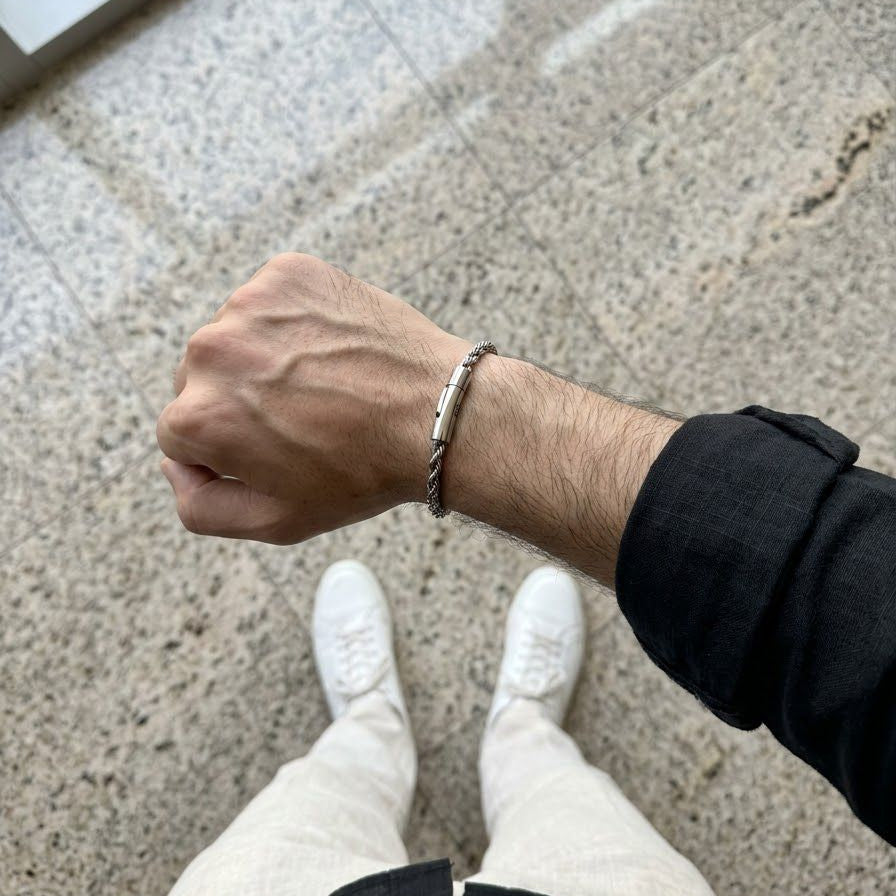 A close-up of a person's forearm and hand wearing a silver-toned twisted rope bracelet, standing on a speckled stone floor with white sneakers and a black shirt visible.