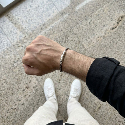 A close-up of a person's forearm and hand wearing a silver-toned twisted rope bracelet, standing on a speckled stone floor with white sneakers and a black shirt visible.