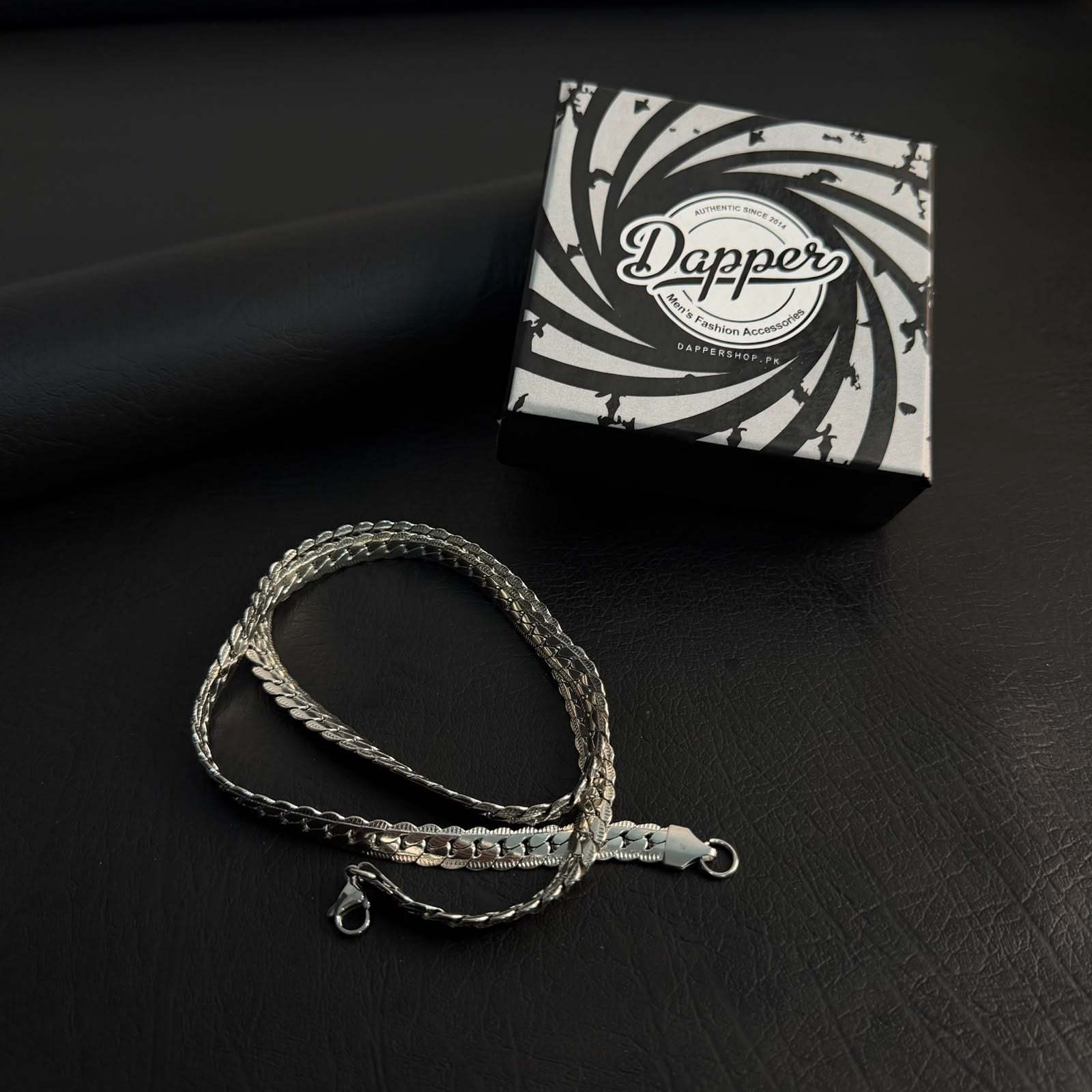 A silver-toned braided chain necklace with a matching metal tag and clasp, displayed on a dark textured surface next to its black and white patterned Dapper brand box.