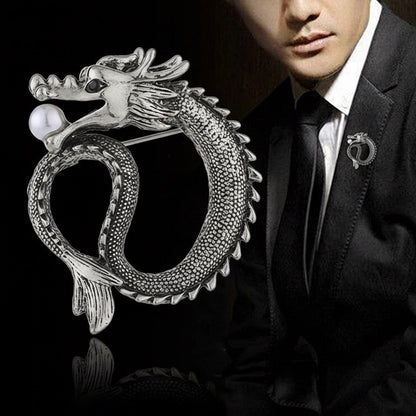 Silver dragon-shaped brooch on a reflective surface with a person wearing a suit in the background.