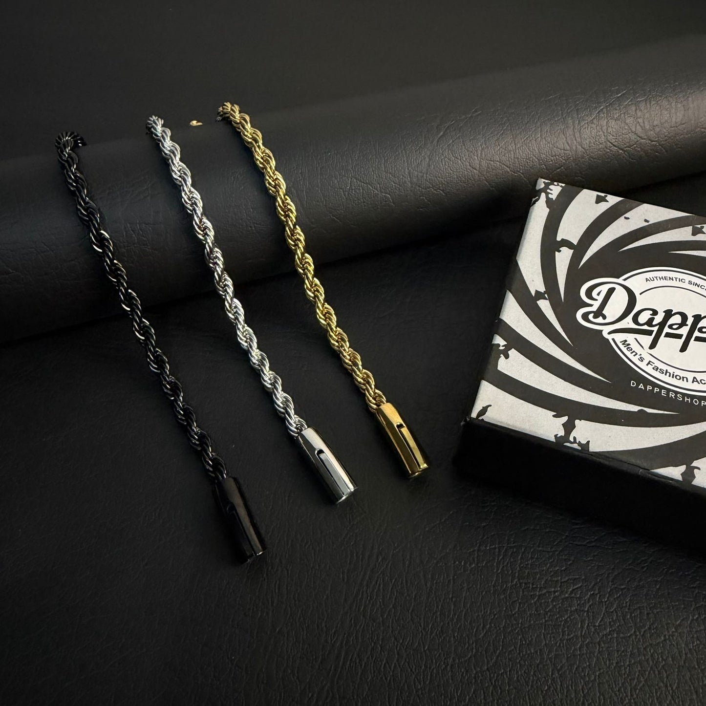 Three rope-style bracelets in black, silver, and gold are displayed on a dark textured surface next to a branded Dappy box.
