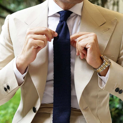 A man wearing a beige suit jacket and trousers, white dress shirt, and a textured navy blue tie, adjusting the tie outdoors with a blurred green garden background.