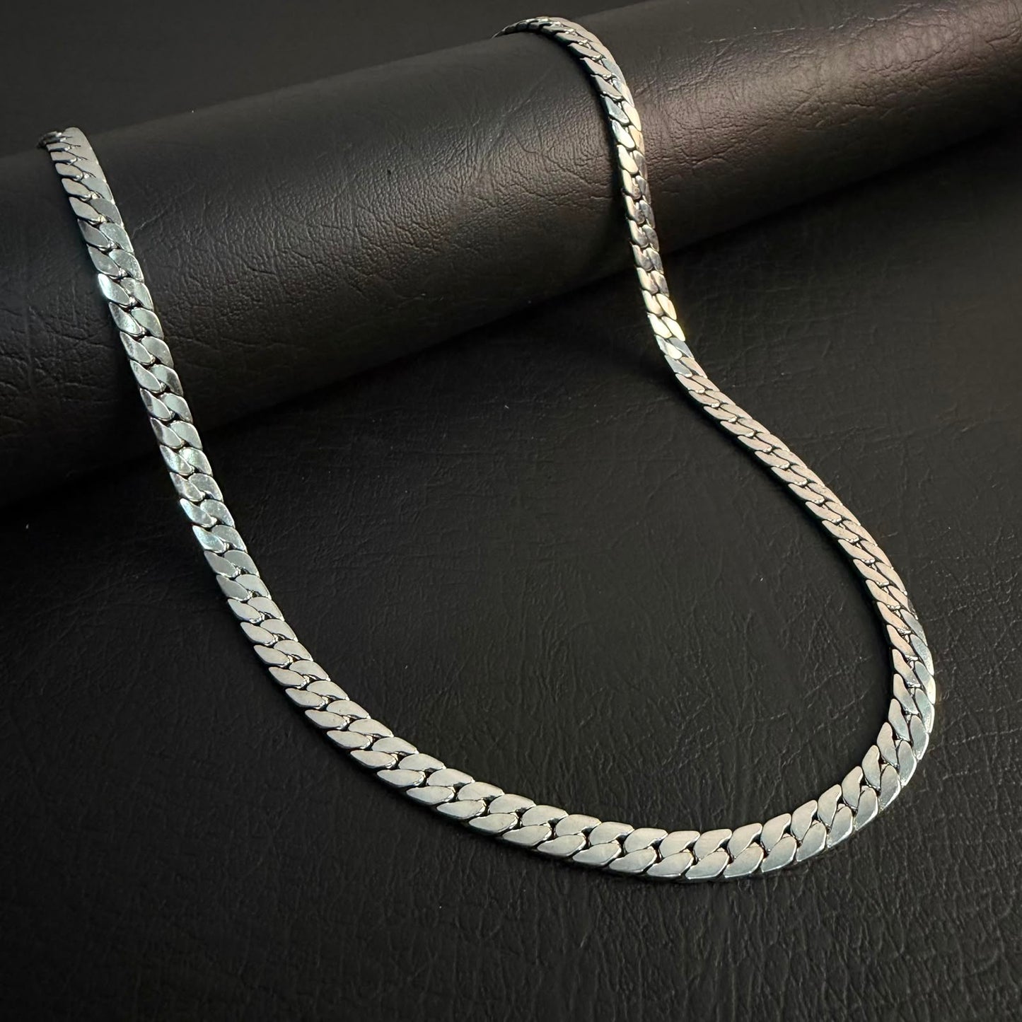 A silver-toned Cuban link chain necklace draped over a black textured cylindrical surface on a dark black background.
