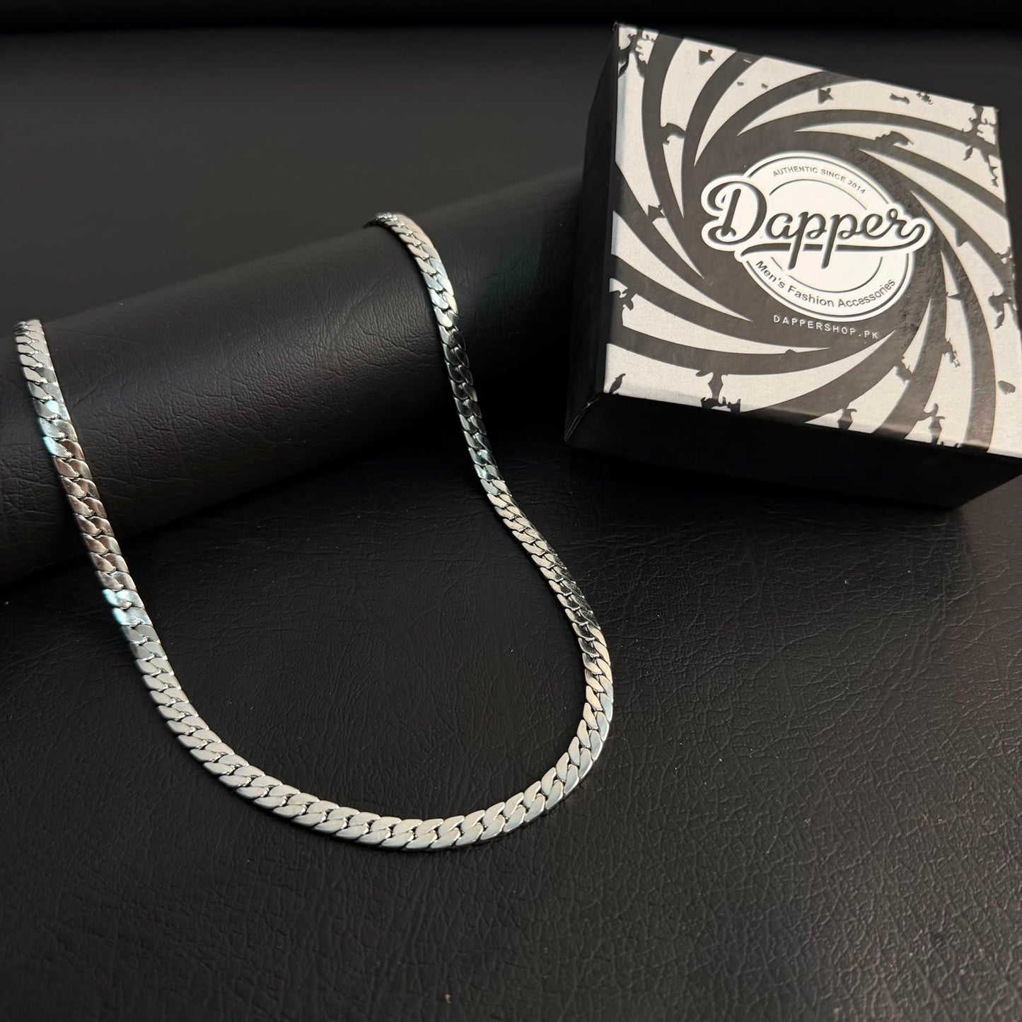 A silver-toned chain necklace draped over a black cylindrical display stand, with a branded black-and-white box labeled 'Dapper' placed behind it on a dark textured surface.