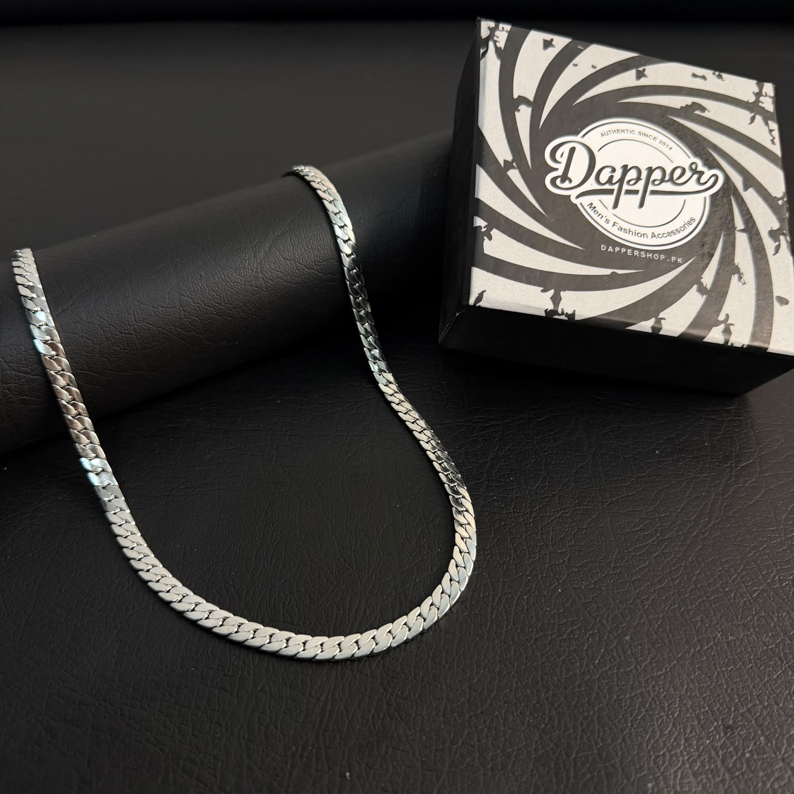 A silver-toned chain necklace draped over a black cylindrical display stand, with a branded black-and-white box labeled 'Dapper' placed behind it on a dark textured surface.