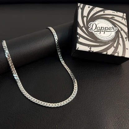A silver-toned chain necklace draped over a black cylindrical display stand, with a branded black-and-white box labeled 'Dapper' placed behind it on a dark textured surface.