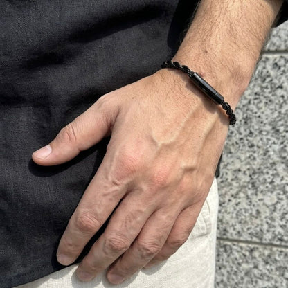 A close-up of a man's hand wearing a black braided cord bracelet with a matte black rectangular metal connector.