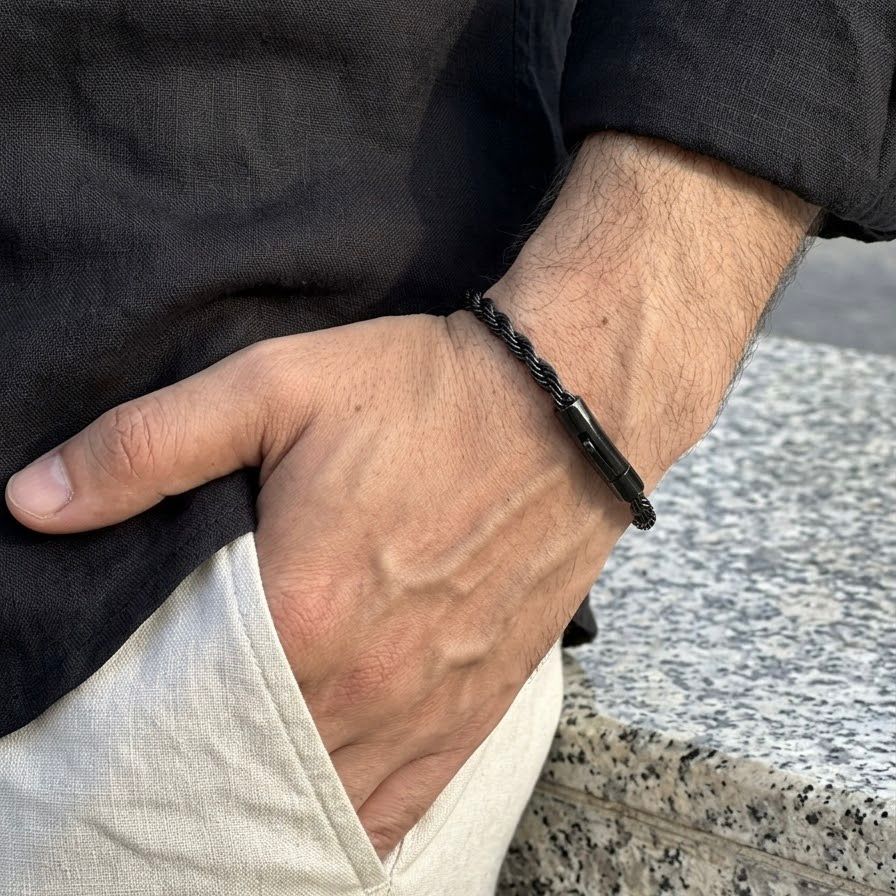 A close-up of a man's hand resting in his pocket, wearing a black braided cord bracelet with a polished black rectangular metal accent piece.