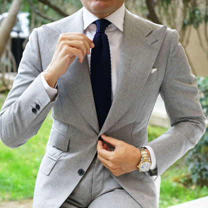 A man wearing a light gray two-piece suit, white dress shirt, and dark navy tie, sitting on a white textured outdoor bench.