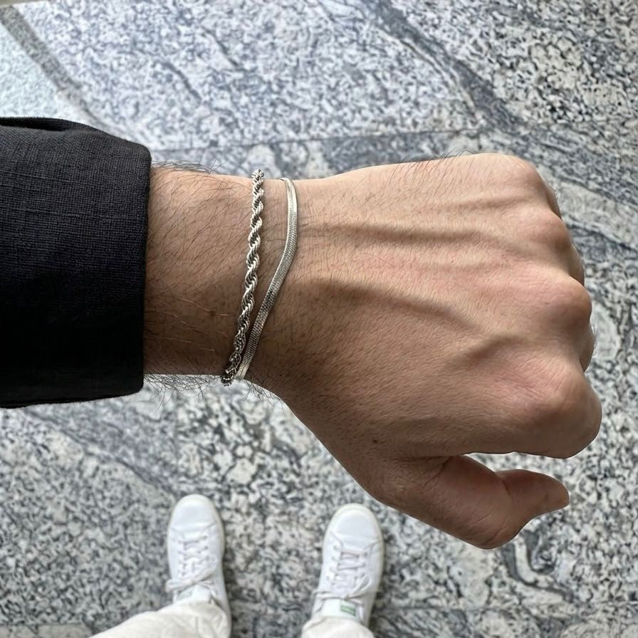 A close-up of a person's wrist wearing two silver bracelets, one a twisted rope-style band and the other a smooth curved bar, standing on a gray stone floor.