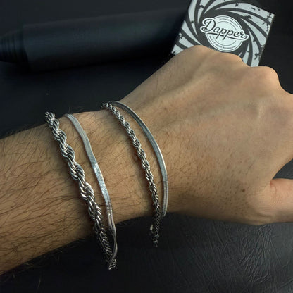 Three silver-toned metal bracelets worn on a person's wrist, including a thick braided rope-style bracelet, a flat snake chain bracelet, and a slim twisted chain bracelet. A black cylindrical item and a black-and-white Dopper branded box are visible in the background.