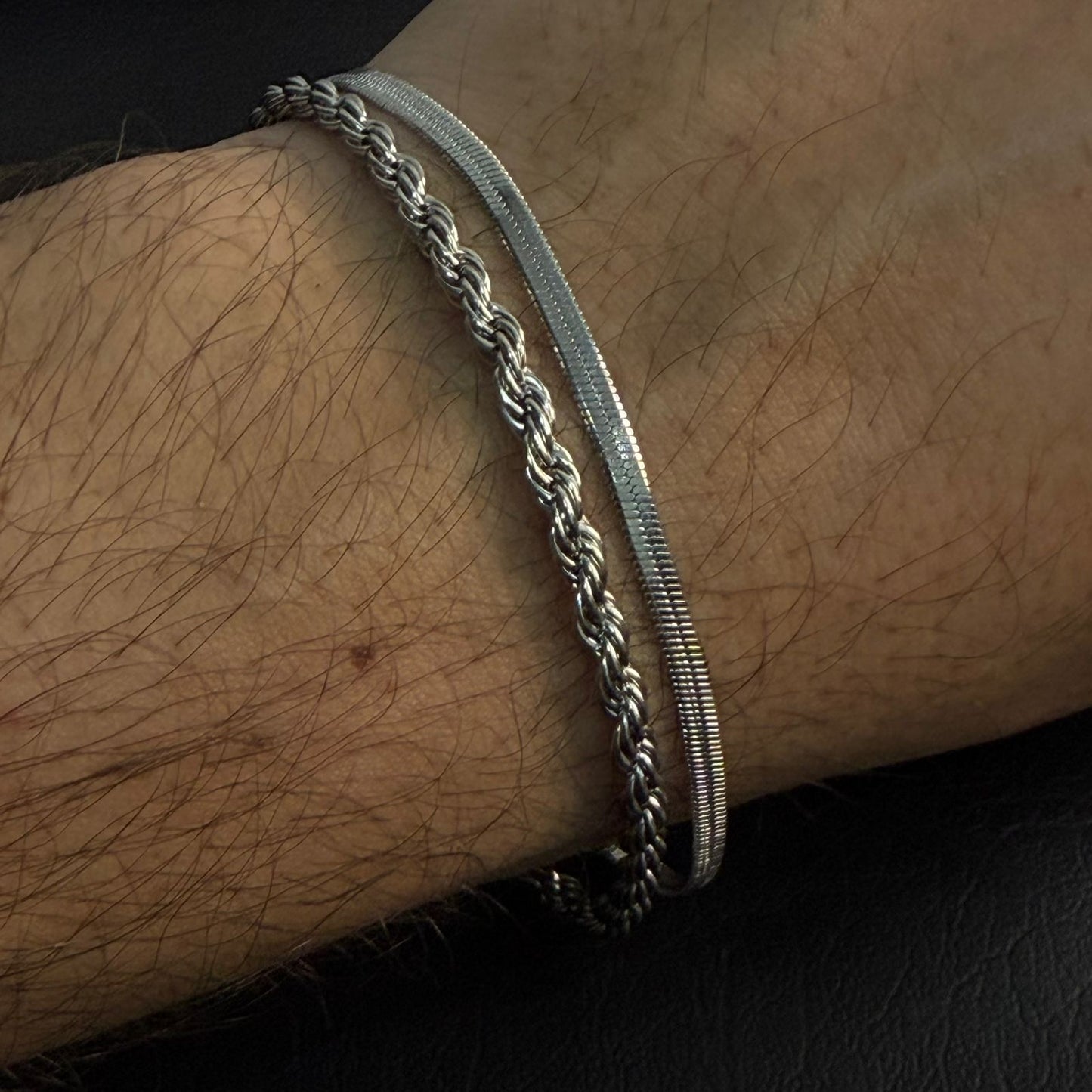 Two silver-toned metal bracelets worn on a hairy wrist, with one thick braided chain and one slim textured snake-style band, against a dark background.