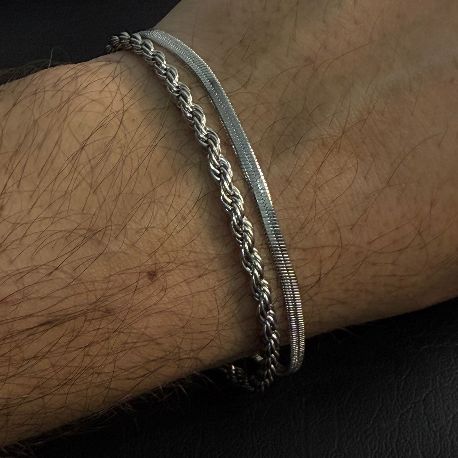Two silver-toned metal bracelets worn on a hairy wrist, with one thick braided chain and one slim textured snake-style band, against a dark background.