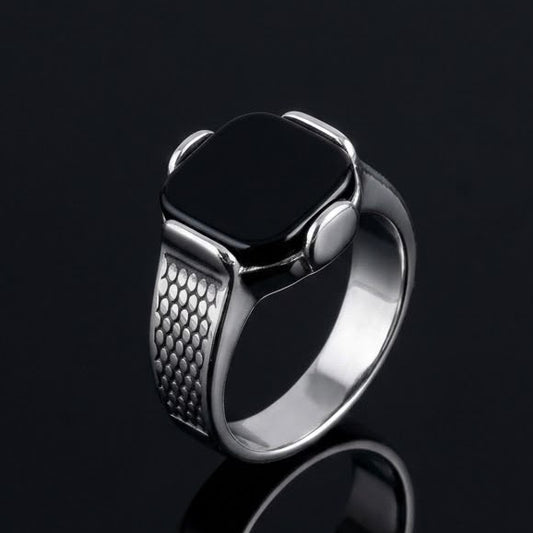 A silver-toned ring with a textured band and a smooth, polished bezel holding a black rectangular inlay, displayed on a reflective black surface.